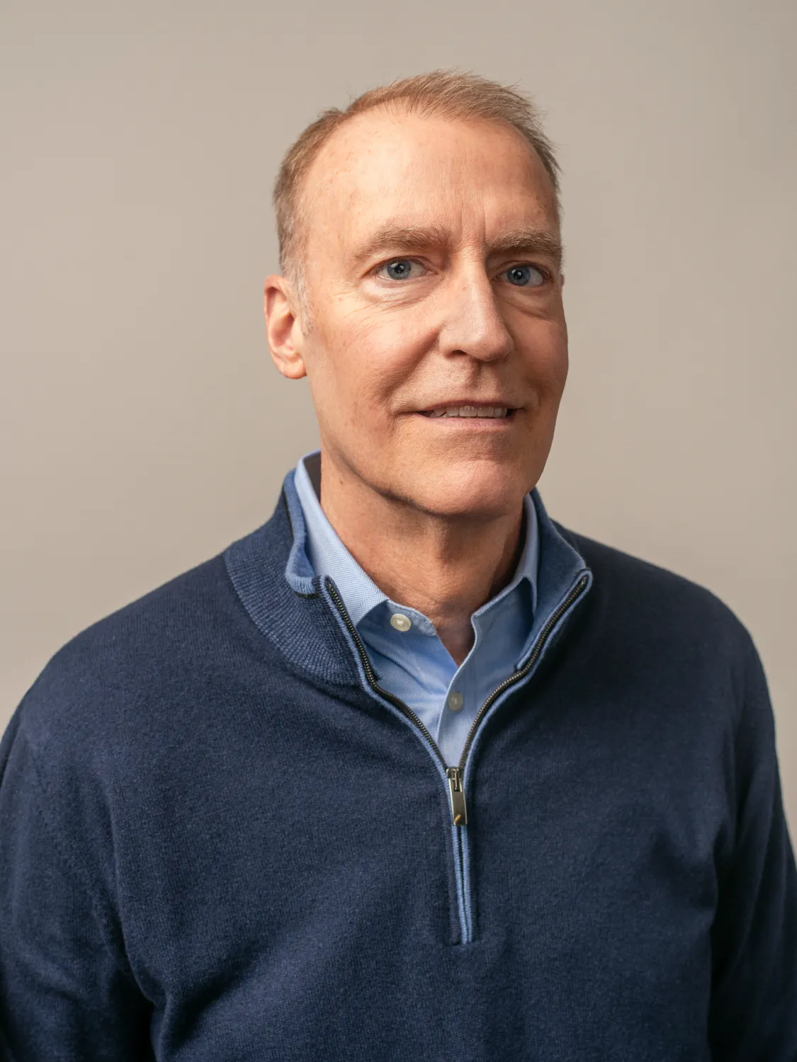Man with short light hair wearing a blue quarter-zip sweater and collared shirt, smiling at the camera against a neutral background.