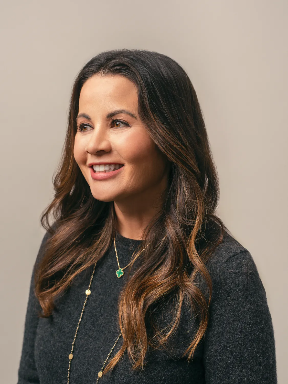 Woman with long brown hair in a dark sweater, smiling and looking to the side against a neutral background.