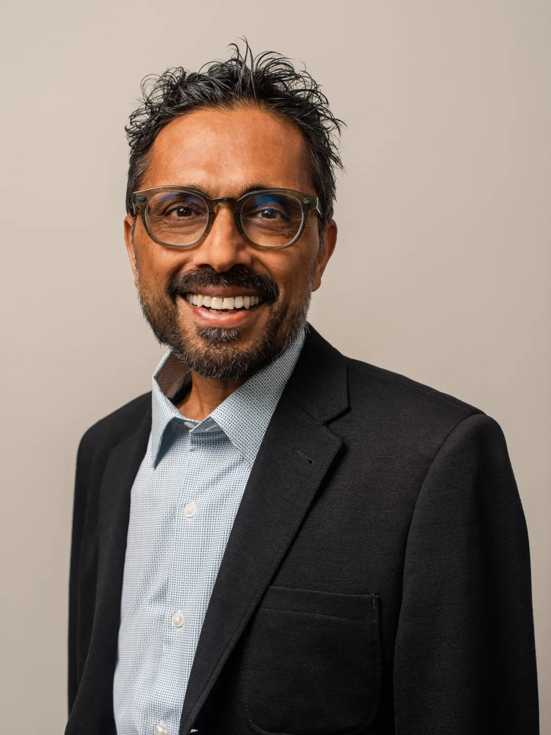 Smiling man with glasses and beard wearing a black blazer and light blue shirt against a neutral background.