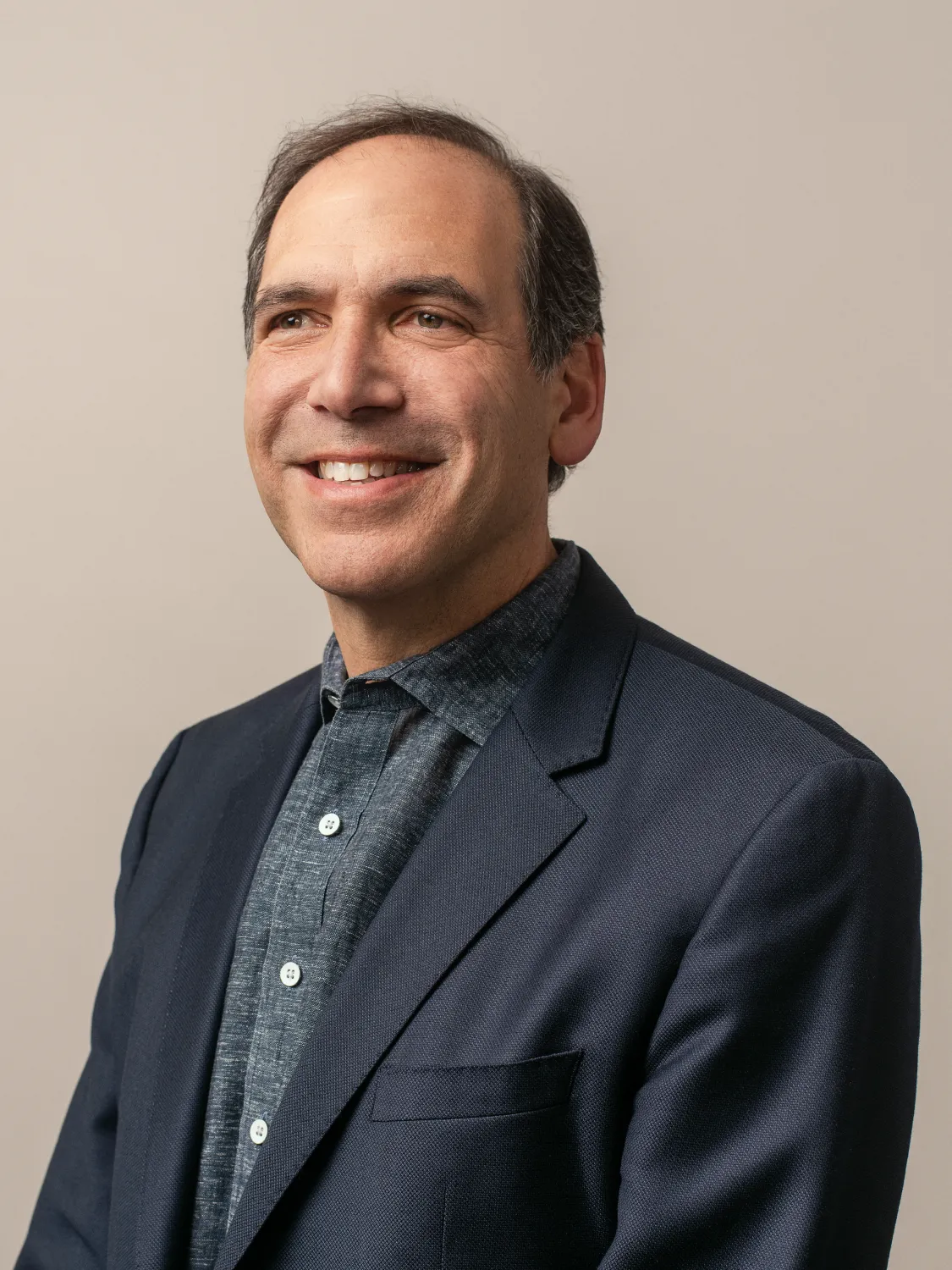 Smiling man in a navy blazer and gray shirt, posing against a neutral background.