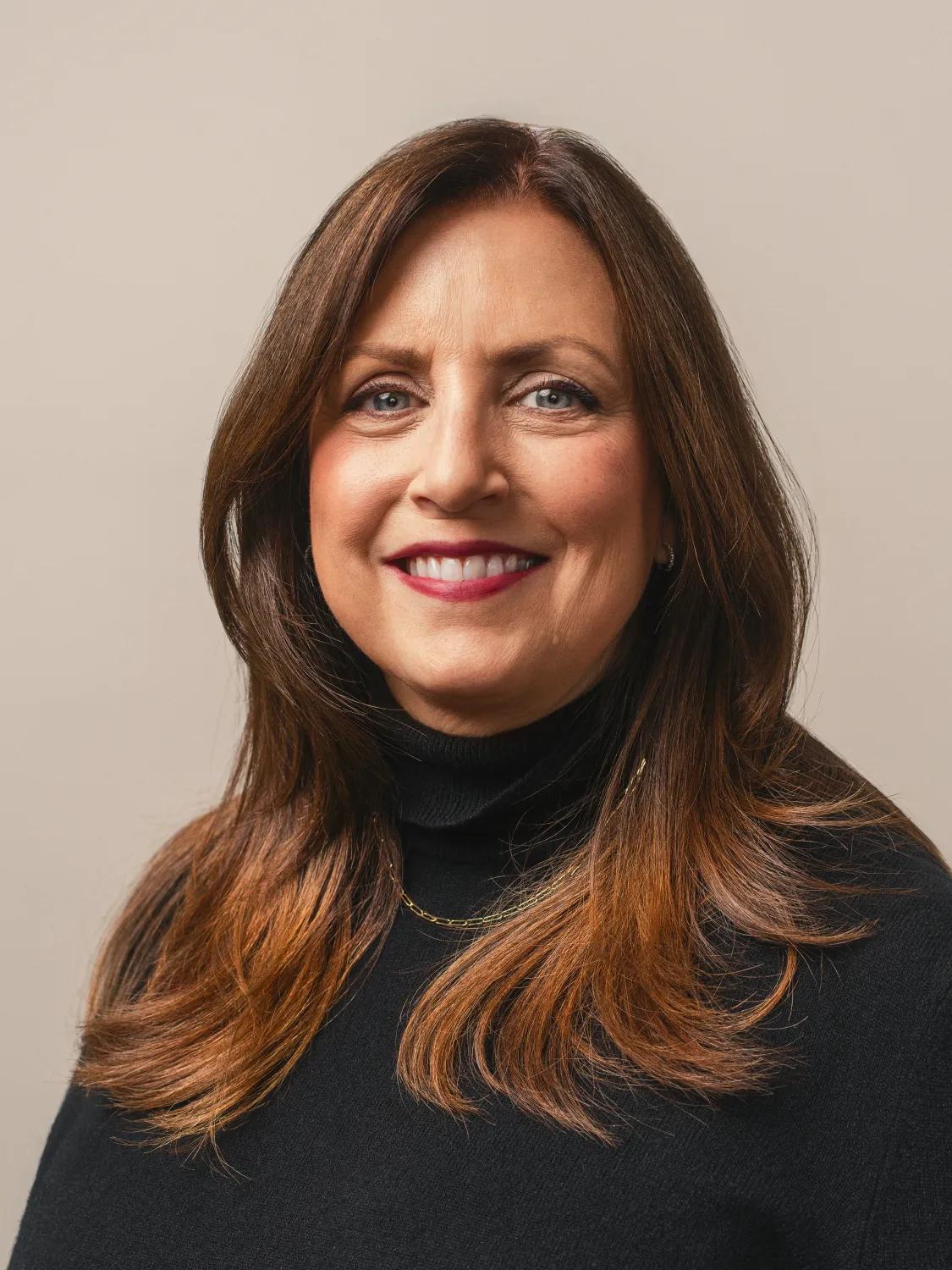Woman with long brown hair, wearing a black sweater, smiling in front of a neutral background.