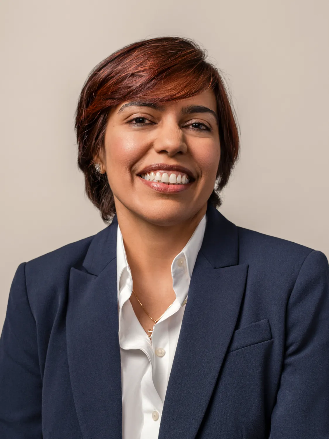 Smiling woman with short brown hair in a navy blazer and white button-up shirt against a neutral background.