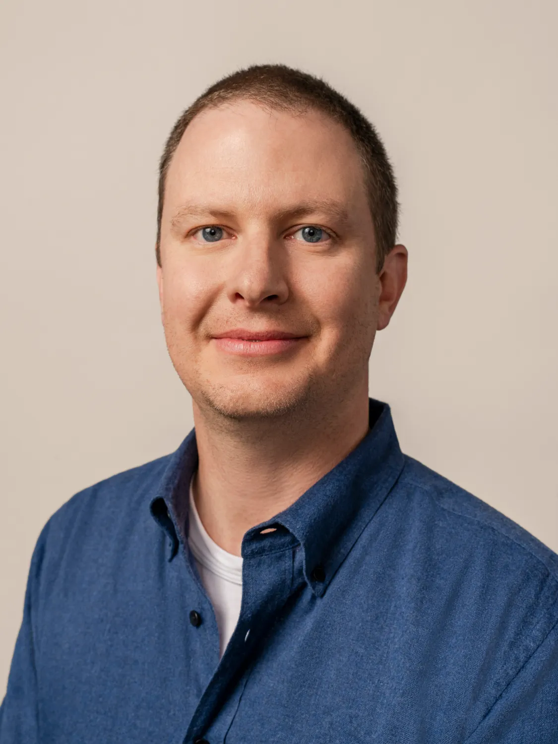 A man with short brown hair wearing a blue button-up shirt and white t-shirt, smiling at the camera on a neutral background.