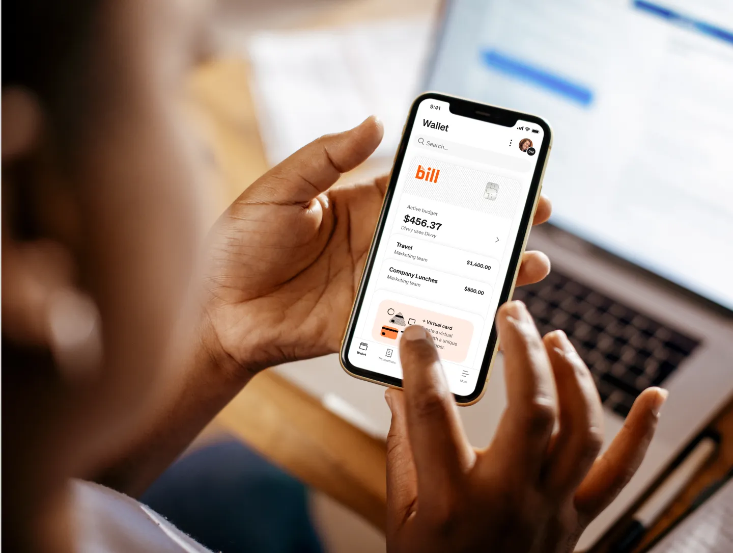 A person uses BILL mobile app on a smartphone showing a digital wallet with active budget of $456.37 while sitting at a desk with a laptop.