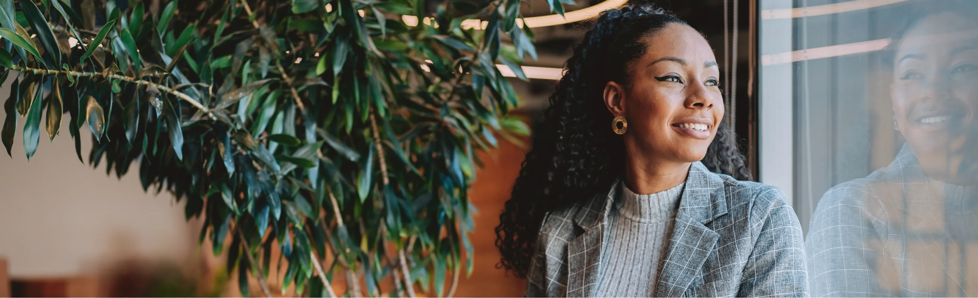 Woman in a gray plaid blazer smiling while looking out a window, with a plant beside her.