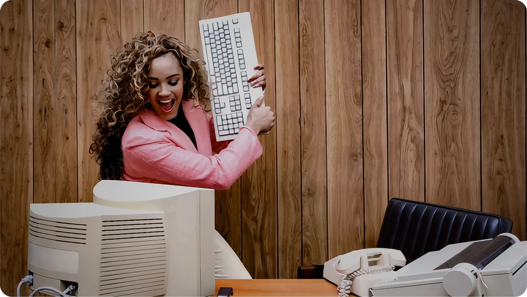 Woman in a pink blazer angrily raises a keyboard over an old computer in a retro office setting.