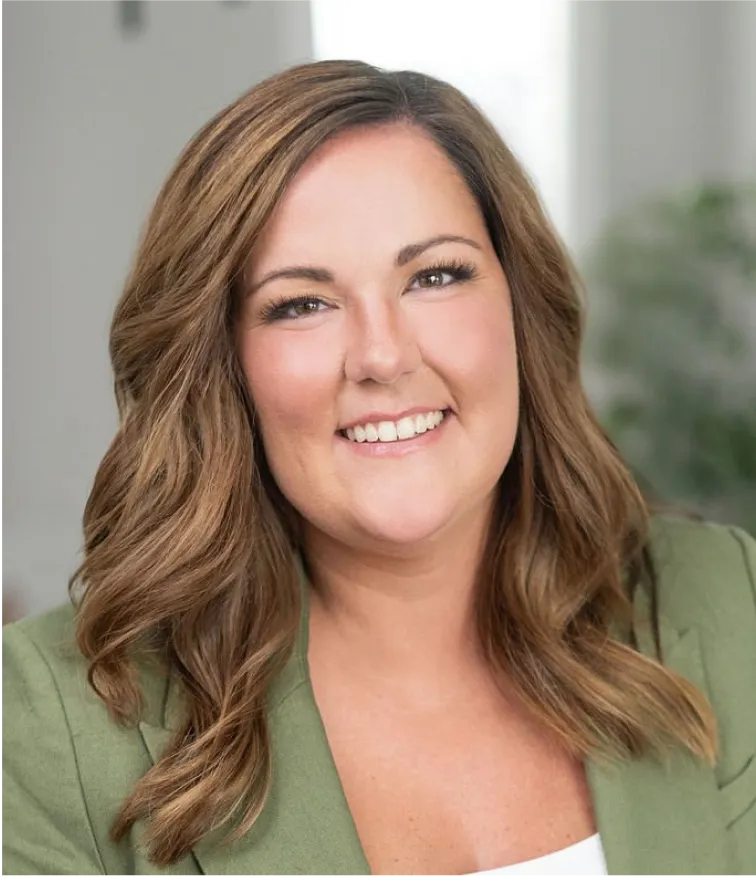 Smiling woman with wavy brown hair wearing a green blazer in a bright indoor setting.