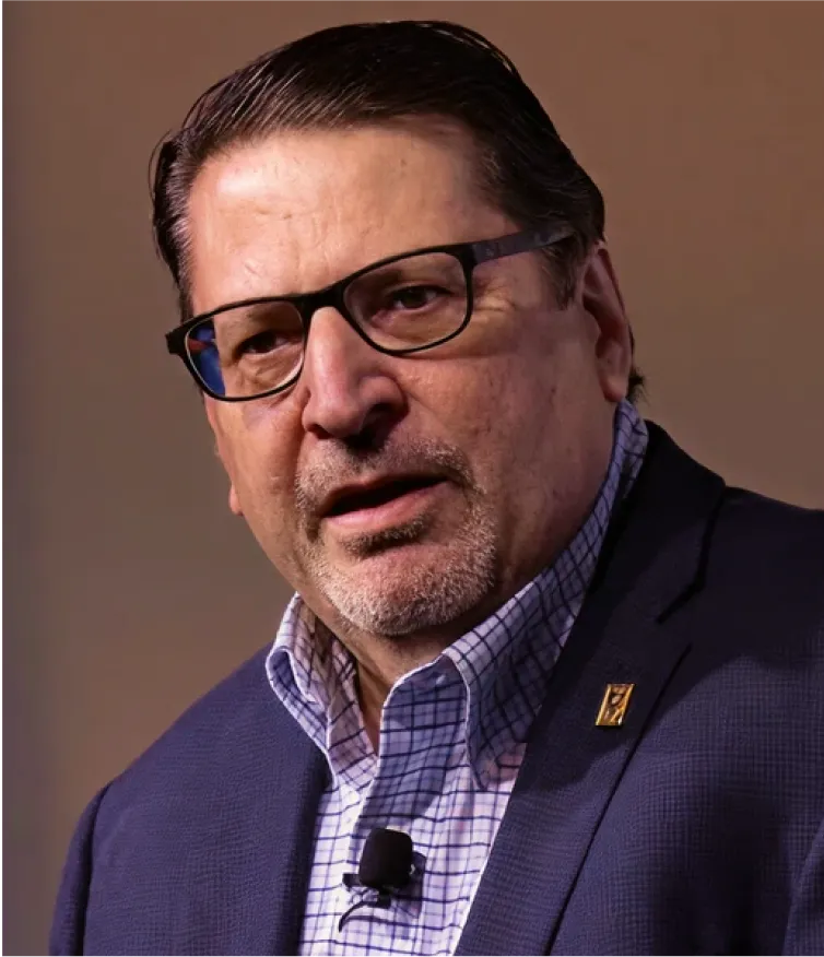 A man in glasses and a suit jacket speaks into a microphone, looking serious.