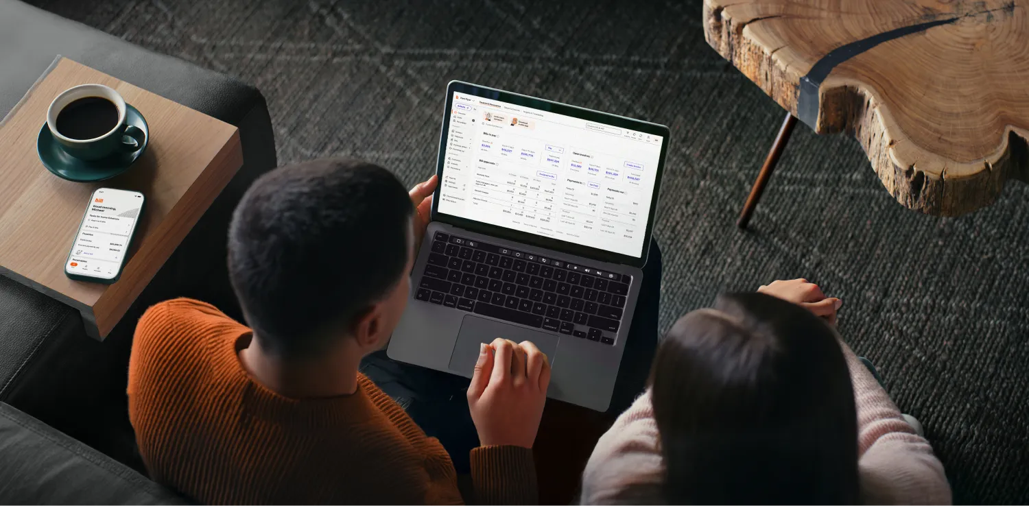 Two people looking at a BILL dashboard on a laptop, with a phone and coffee cup on a nearby table.