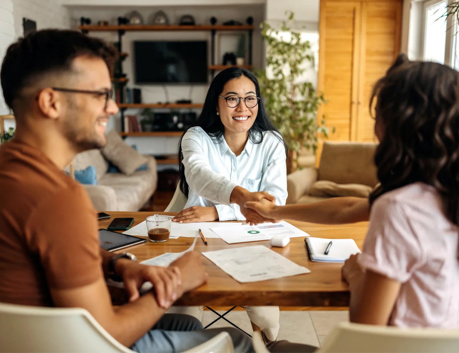 A smiling woman shakes hands with a client across a table with papers and coffee cups.