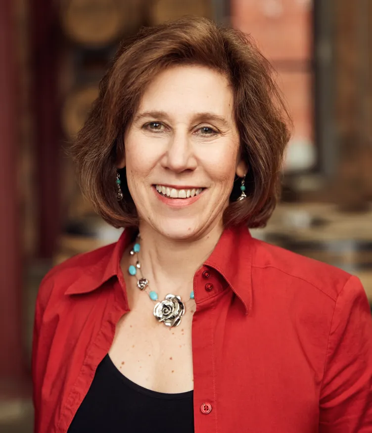 Woman with brown hair wearing a red shirt and a flower necklace, smiling in an indoor setting.