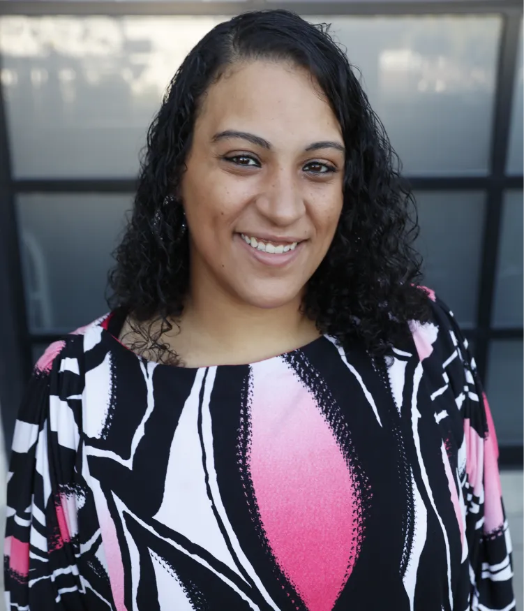 Woman with curly hair smiles, wearing a pink, white, and black patterned top, standing in front of glass doors.