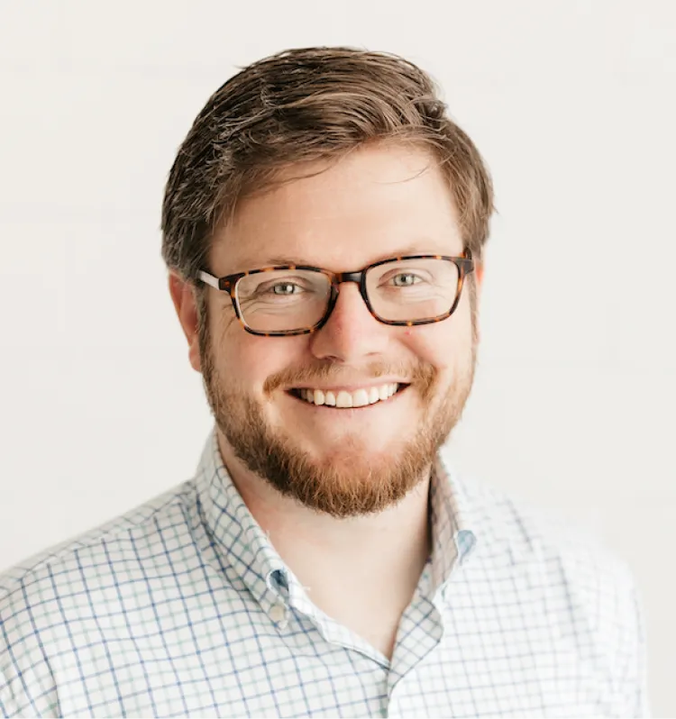 Smiling man with glasses, short brown hair, and a beard, wearing a checkered shirt, against a white background.