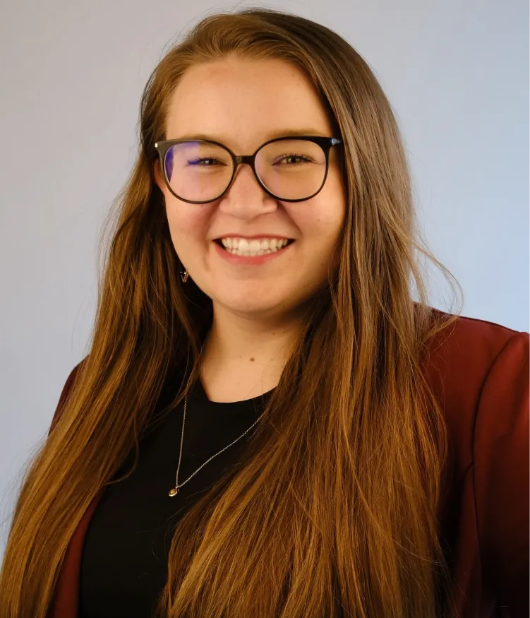 Woman with long brown hair, glasses, and a maroon blazer smiling against a plain background.