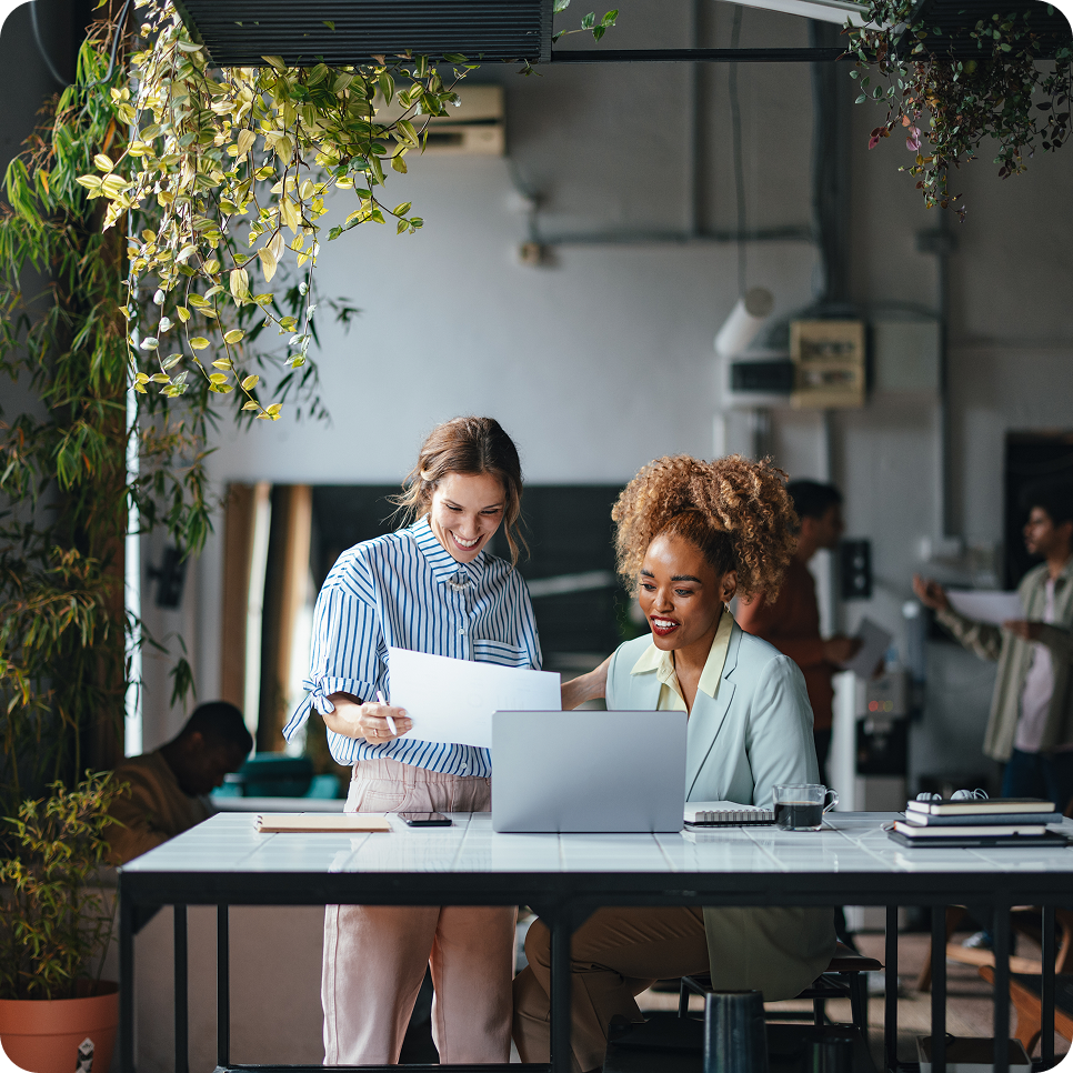 Two women smiling and reviewing documents together at a desk in a modern office with plants.