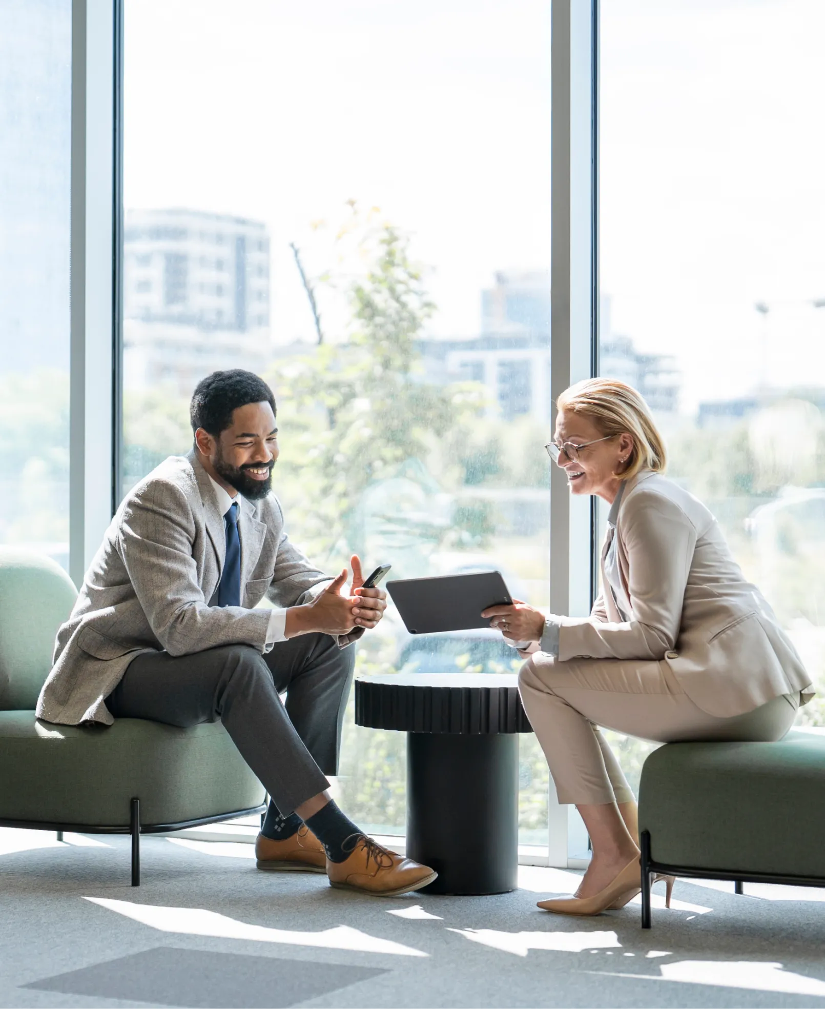 Two businesspeople sit across from each other, smiling, talking, looking at a tablet in a modern office with large windows.