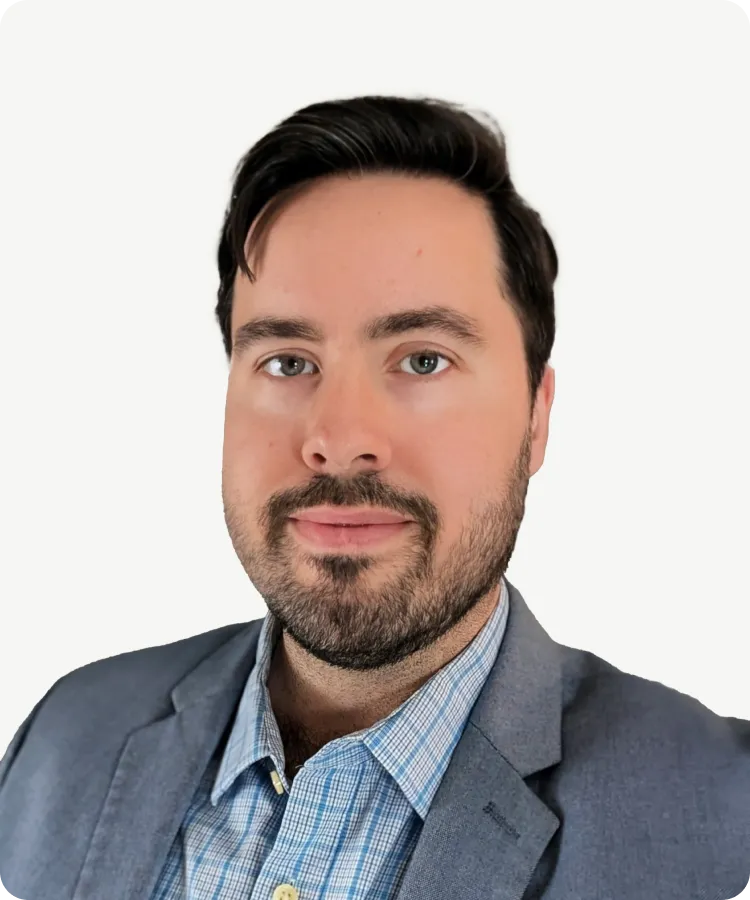 Man with dark hair and beard, wearing a gray blazer and checkered shirt, posing against a white background.