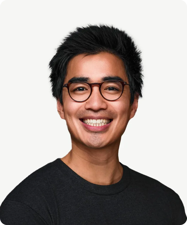 A man with short black hair and glasses smiles, wearing a black shirt, against a plain white background.