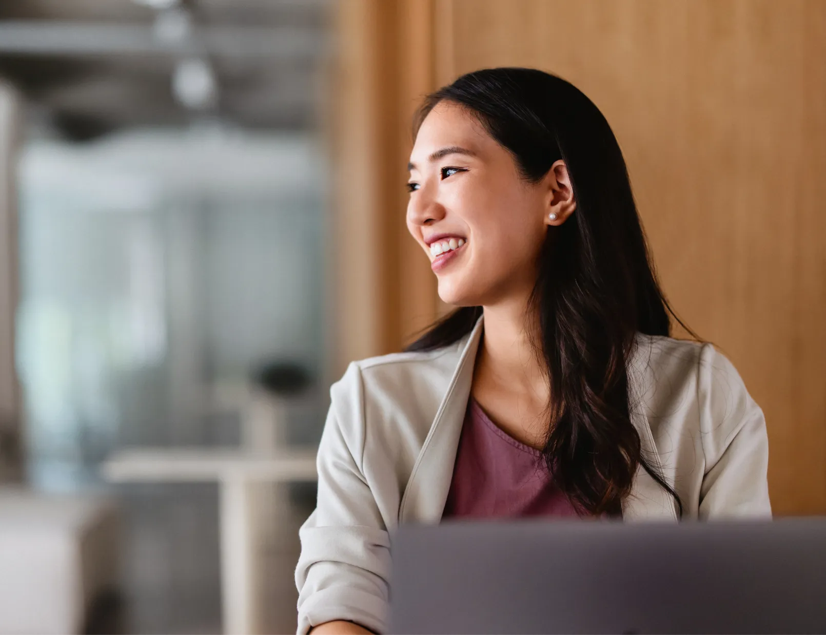 Woman smiling and looking to the side while sitting at a desk with a laptop in a modern office.