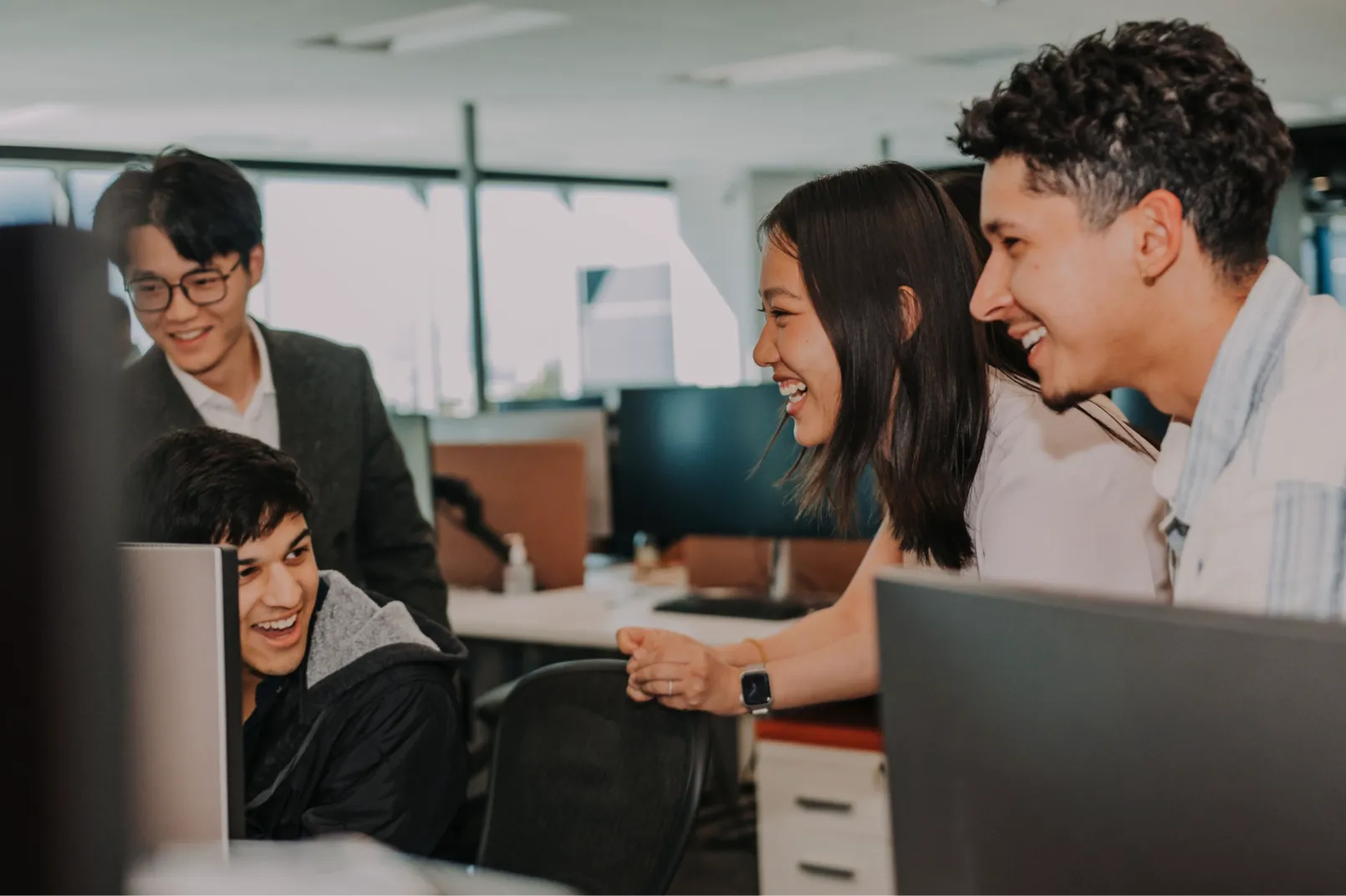 Four young adults laughing and collaborating around computer screens in a bright office setting.