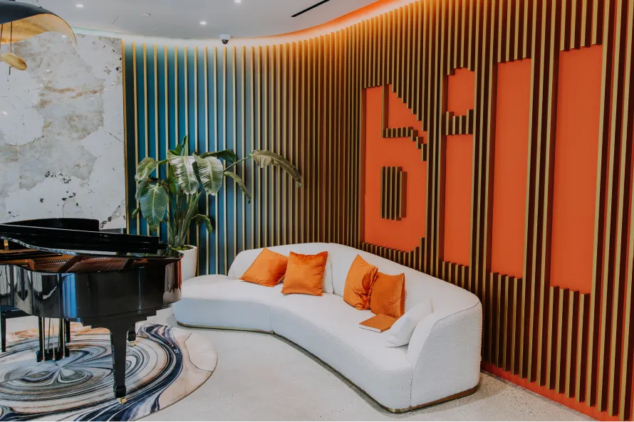 Modern office lobby with a black grand piano, white curved sofa, orange pillows, and wooden stripes wall decor with "bill" embedded in orange.
