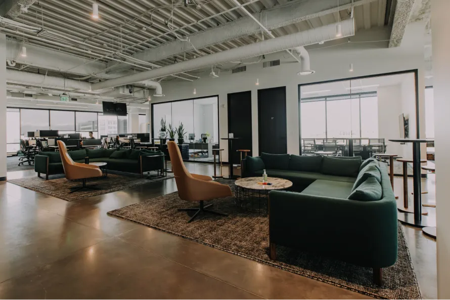 Modern office lounge with green sofas, armchairs, coffee tables, and large windows in the background.