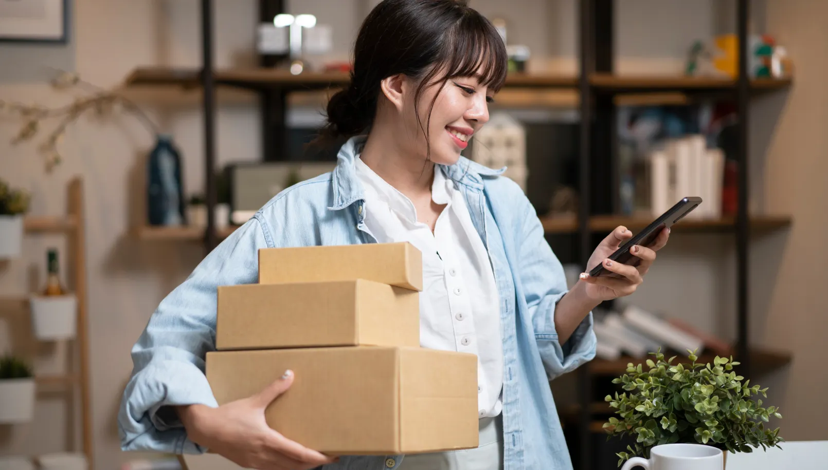 Smiling woman holding cardboard boxes looks at her phone in a home office setting.