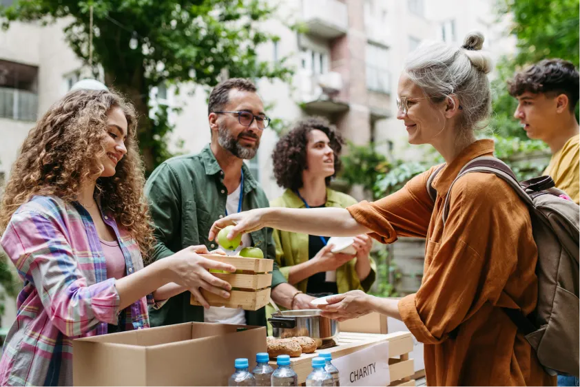 People handing out food and drinks at an outdoor charity event with trees and buildings in the background.