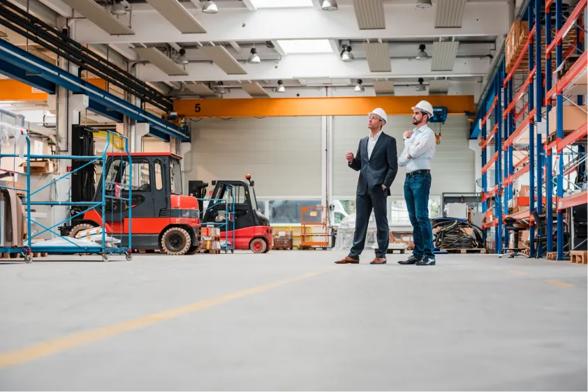 Two men in hard hats stand in a spacious warehouse with shelves and forklift in the background.