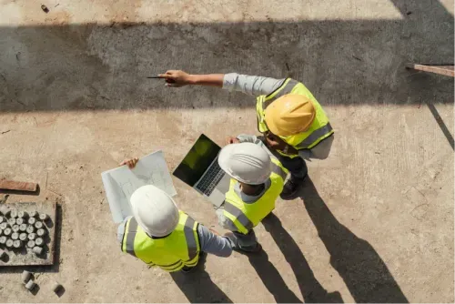 Three construction workers in safety gear review blueprints and a laptop on a construction site, viewed from above.