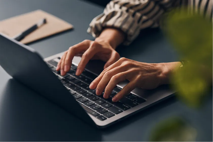 Close-up of hands typing on a laptop keyboard, with a notepad and pen in the background.