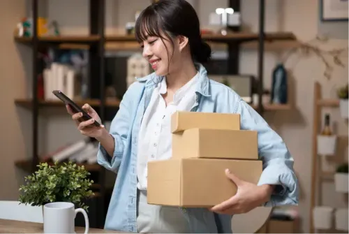 Smiling woman holding cardboard boxes and looking at her phone in a home office setting.