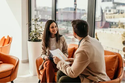 Two people in business attire sit on orange chairs, smiling and talking, holding paperwork in a bright office.