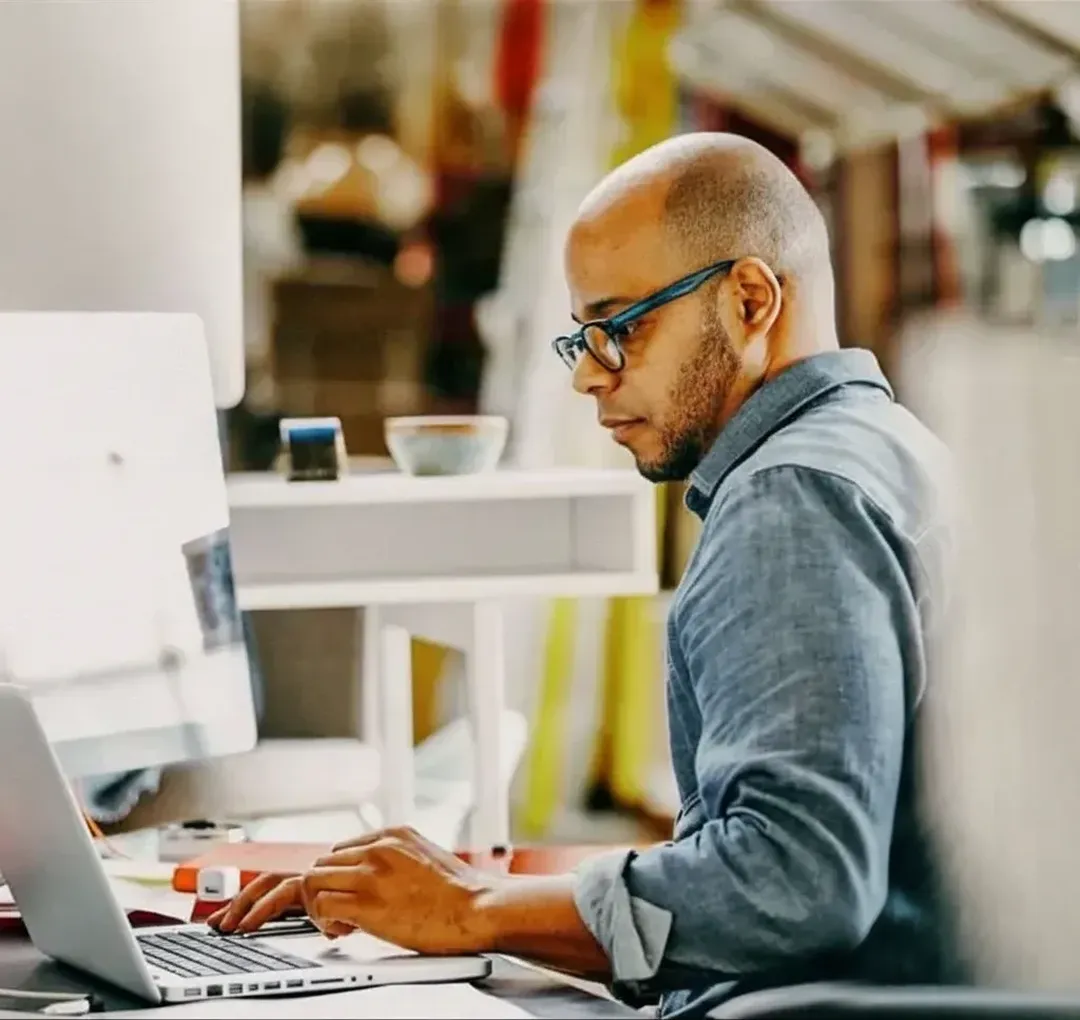 A man wearing glasses and a denim shirt works at a laptop in a bright, modern office.
