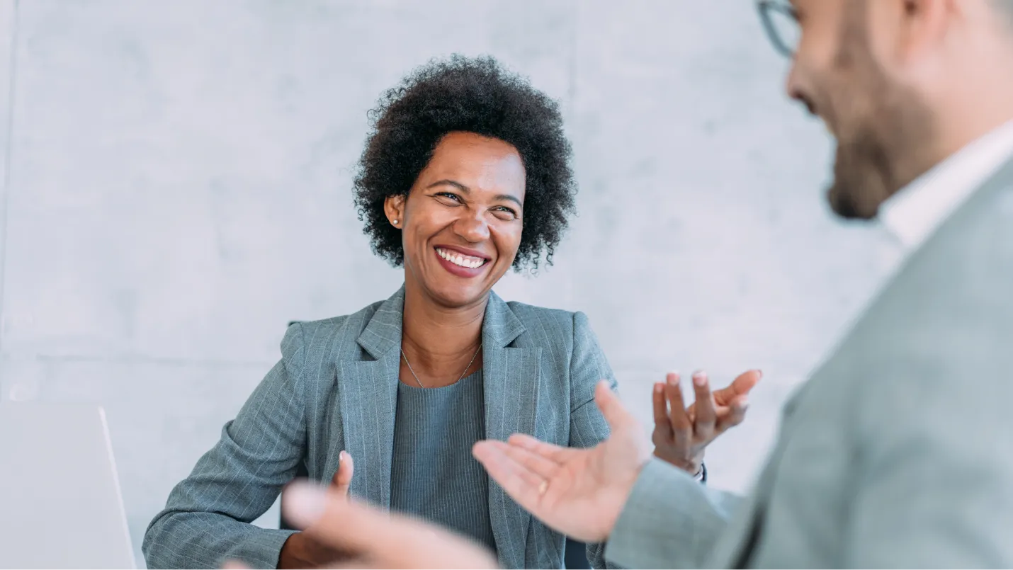 Laughing woman has a conversation with a smiling man in a white, modern room, a laptop sits on a table beside the woman.