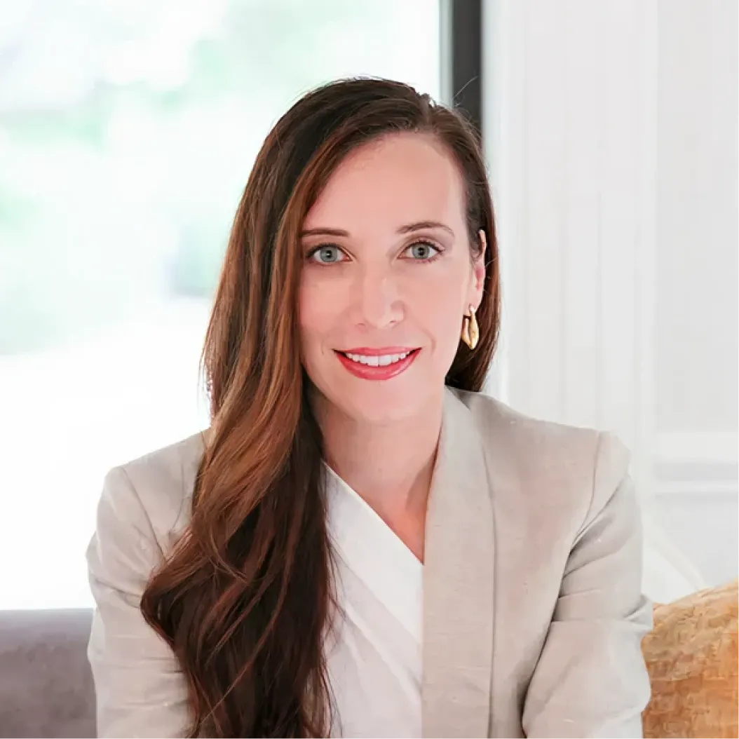 Smiling woman with long brown hair wearing a beige suit and white blouse sits on a sofa in front of large windows.