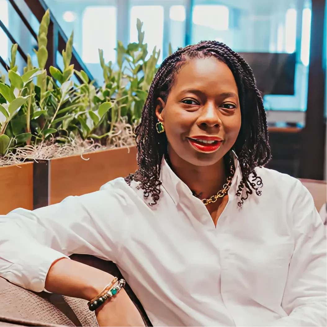 Woman in a white blouse smiles while leaning one arm on the back of a sofa in an office setting, plants and large windows are in the background.