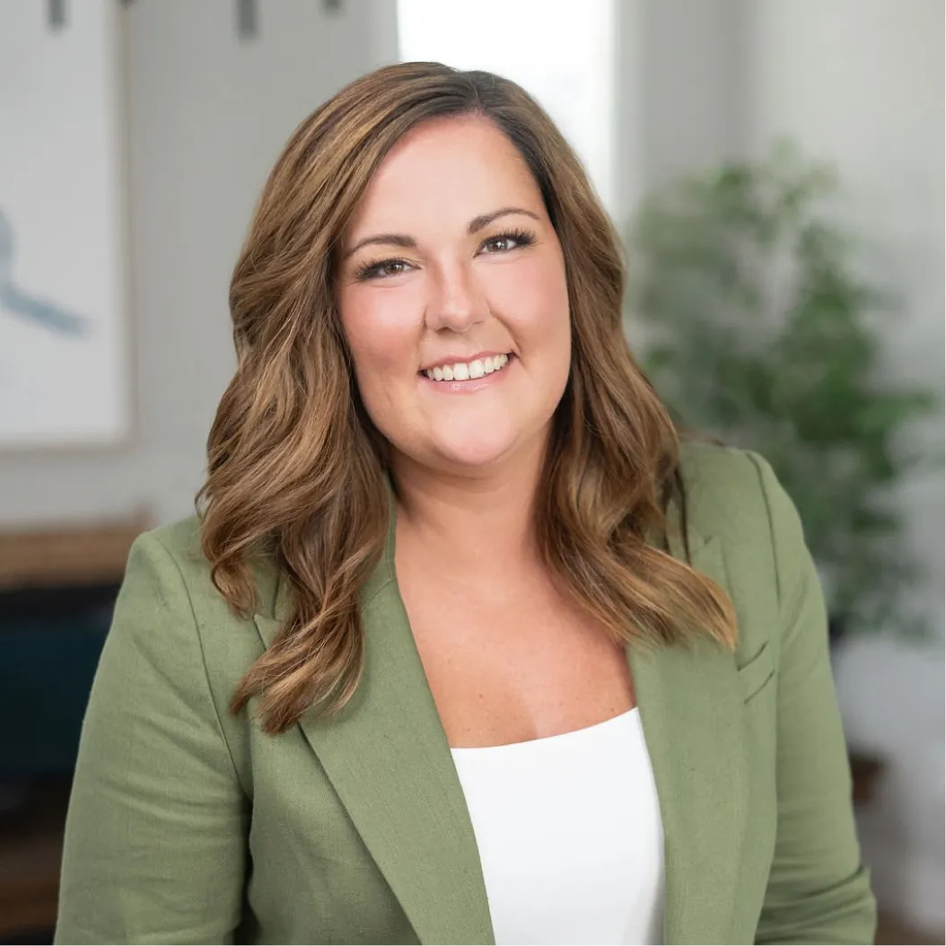 Smiling woman with light brown long hair in an olive green blazer and white shirt sits in a modern office setting with a plant and windows in the background.