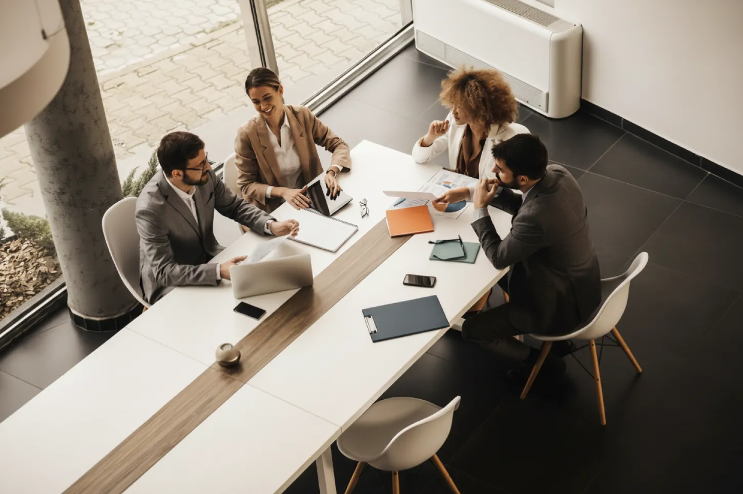 Four professionally dressed men and women sit at large white conference table beside large windows.