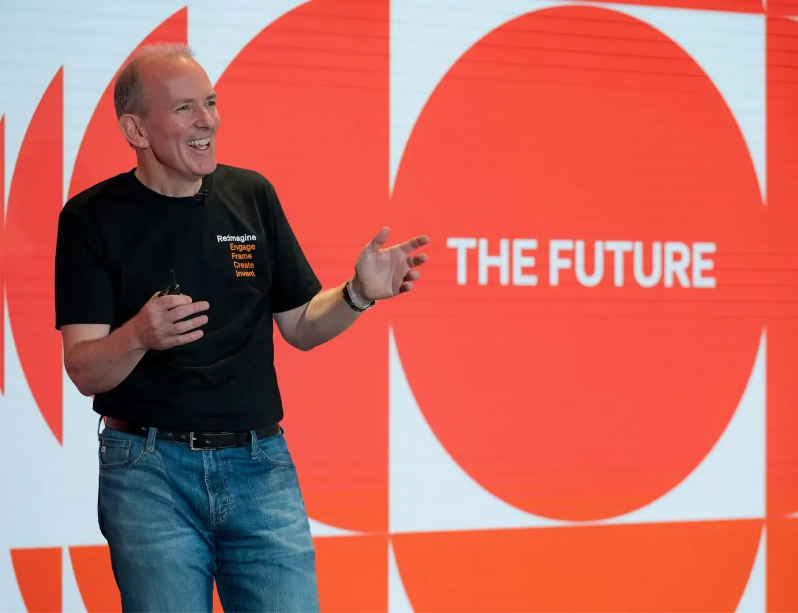 BILL founder and CEO, René Lacerte, wearing a black BILL t-shirt and blue denim jeans, presenting on stage in front of a graphic orange and white backdrop with "The Future" in white text.