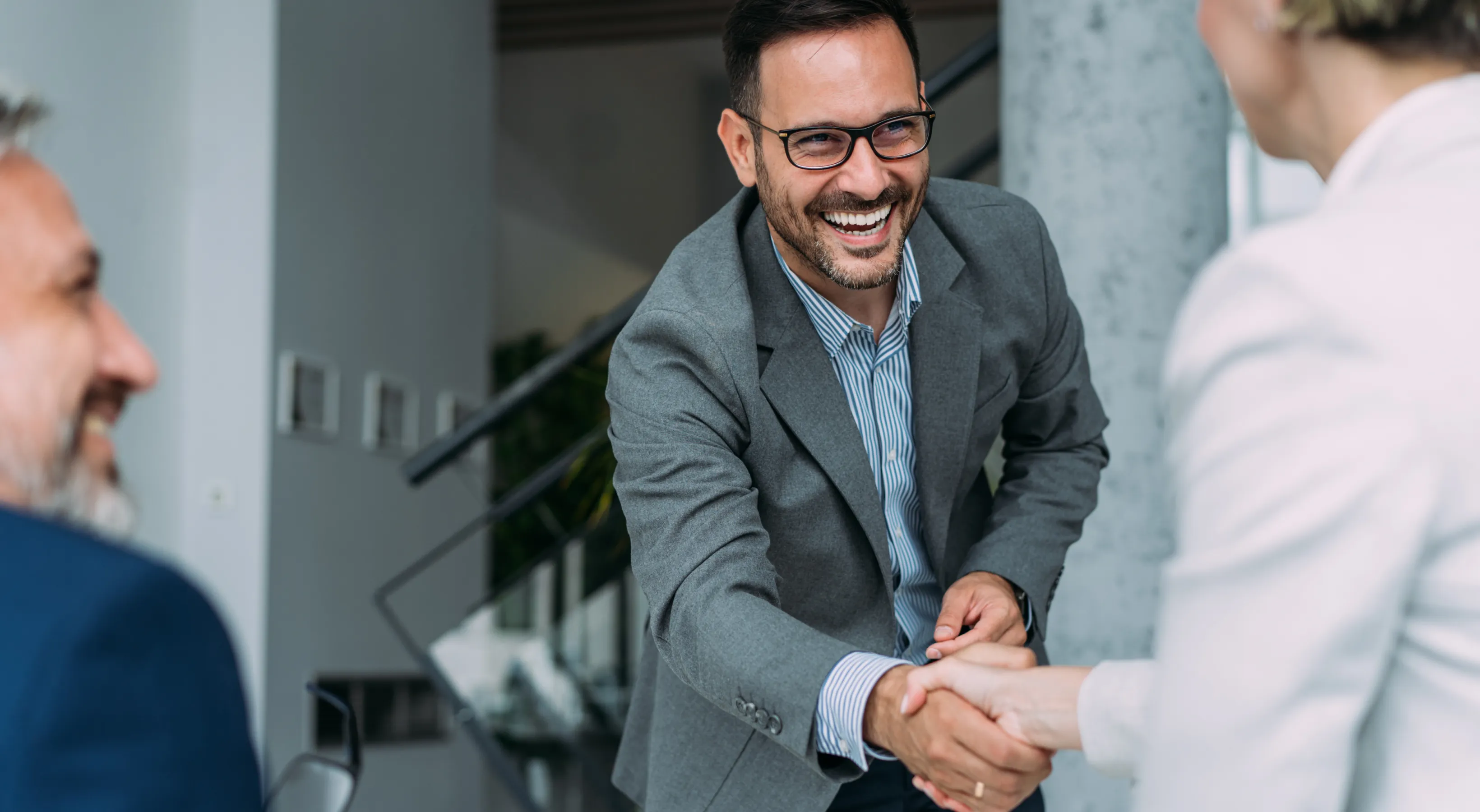 Smiling man with glasses wearing a light blue shirt and grey blazer stands in a modern office, reaches across a table to shake hands with two people.