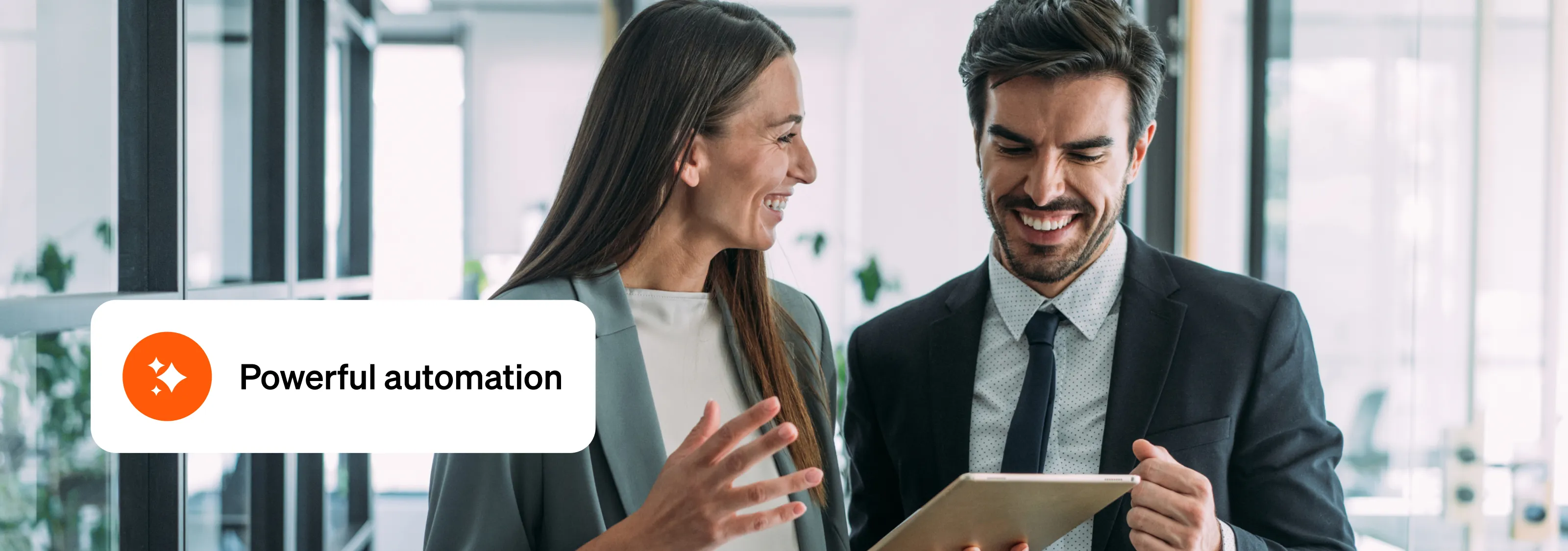 Smiling man and woman in professional attire have a conversation, holding a tablet, in a bright office setting, an overlay reads "Powerful automation".