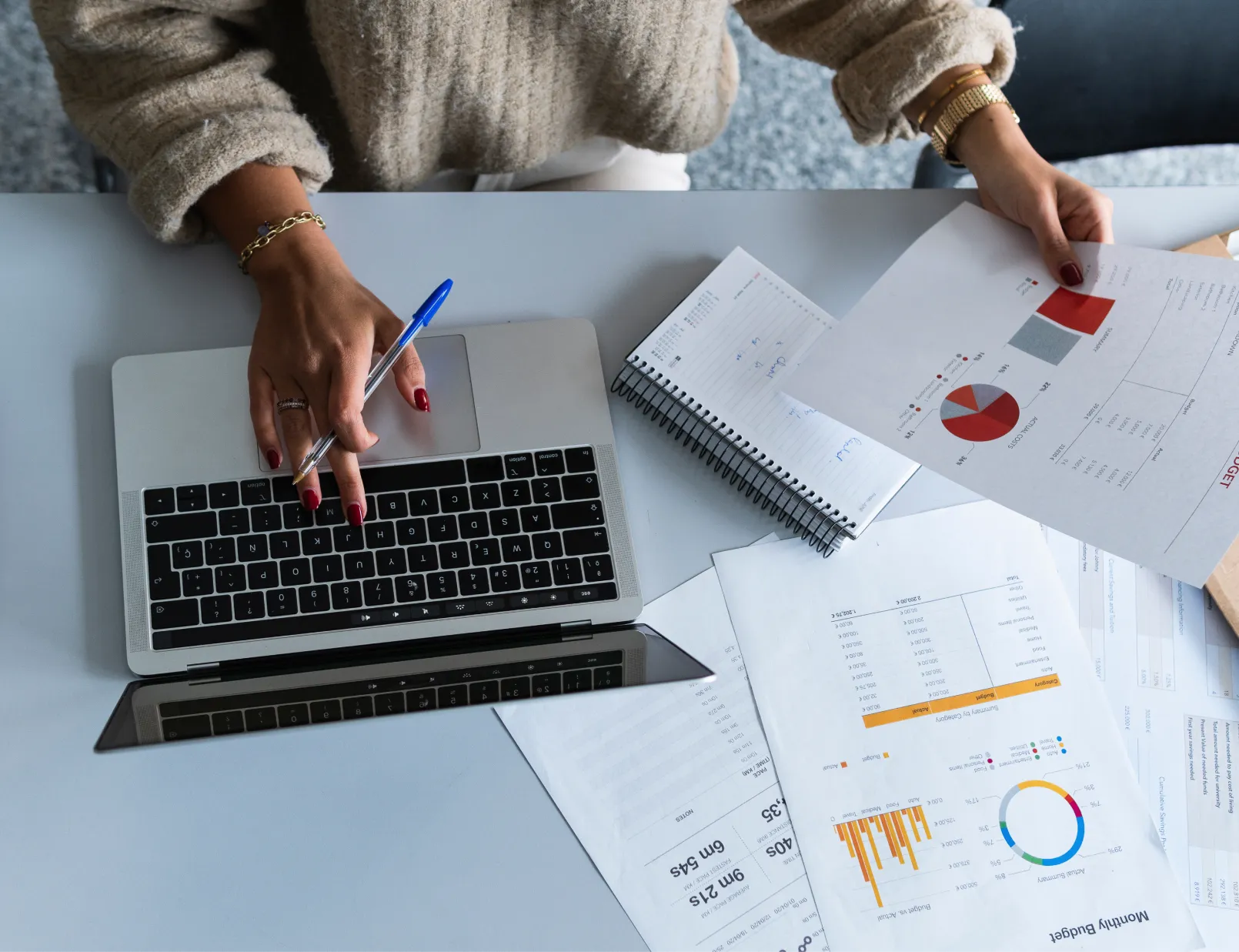 A person works on a laptop, papers with charts and graphs lay on a modern white table.