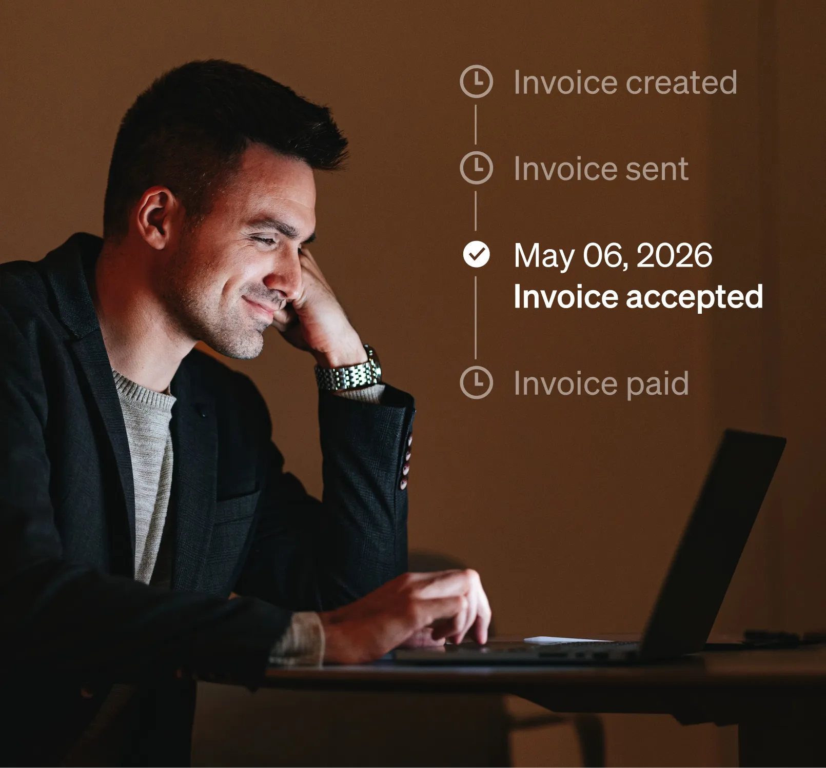 Man in a light grey sweater and dark blazer sitting at a table working on a laptop in a low lit office, laptop notifications show Invoice created and Invoice sent completed, invoice accepted in progress.
