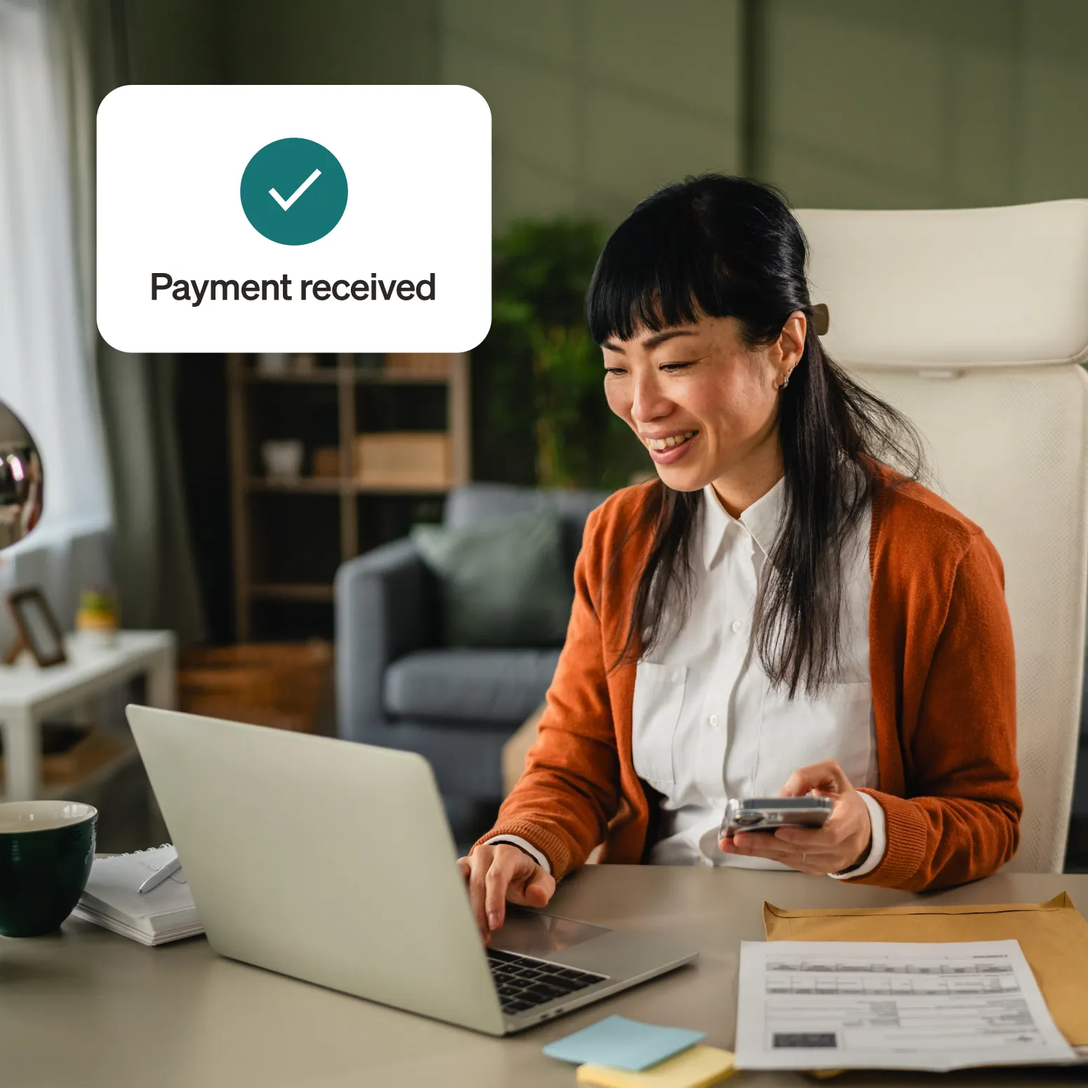 Smiling woman sits at a desk in an executive office working on a laptop showing a notification "Payment received" and a green checkmark, papers and post-it notepads are on the desk around her.
