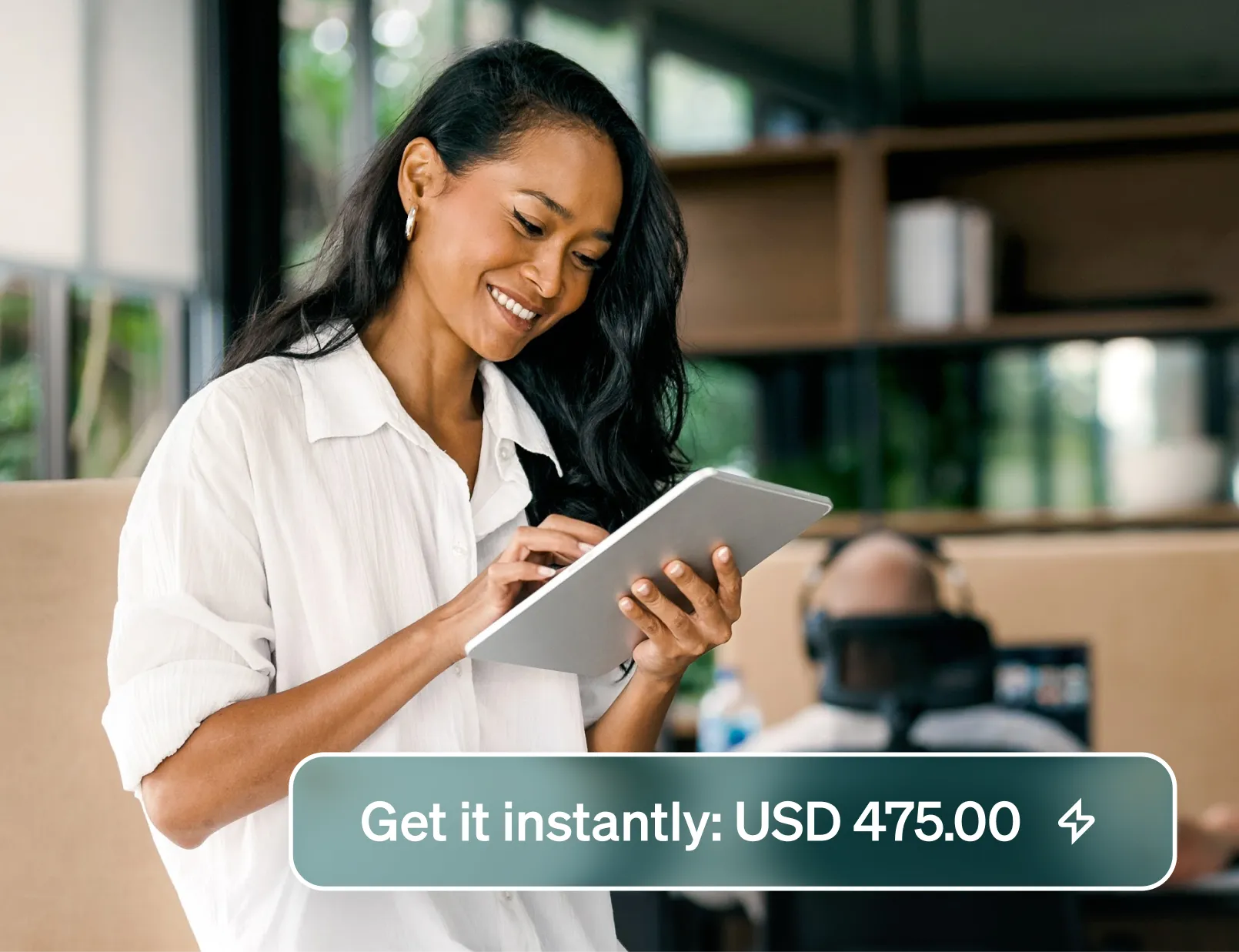 Smiling woman in a bright office with large windows uses a tablet showing a notification "Get it instantly USD 475.00".