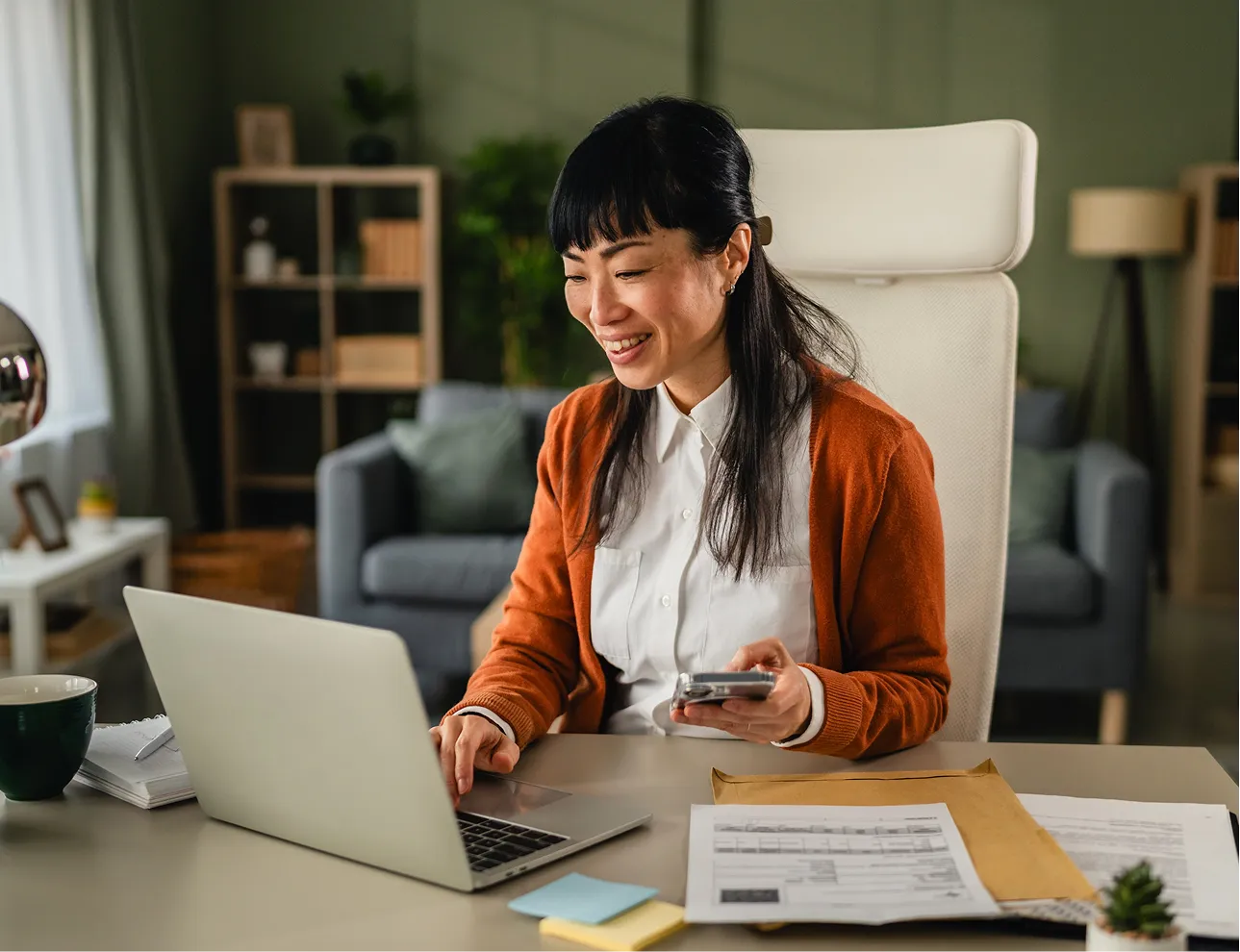 Smiling woman in a large office working on a laptop at a desk, papers and post-it notes on the desktop.