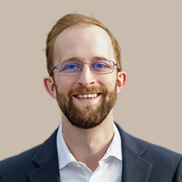 Man with red hair and beard wearing glasses, a white shirt, and a dark blazer smiles in front of a neutral background.