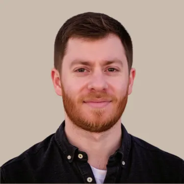 Man with dark hair and short brown beard wearing a dark blue button-down shirt in front of a neutral background.