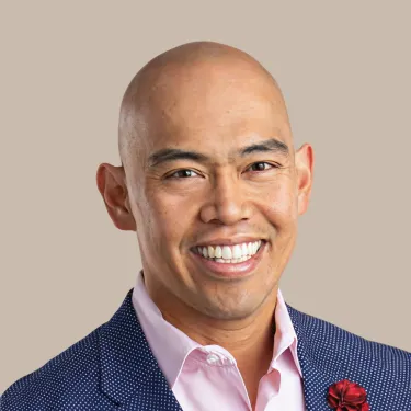 Bald man wearing a pink shirt and dark blue jacket smiles in front of a neutral background.